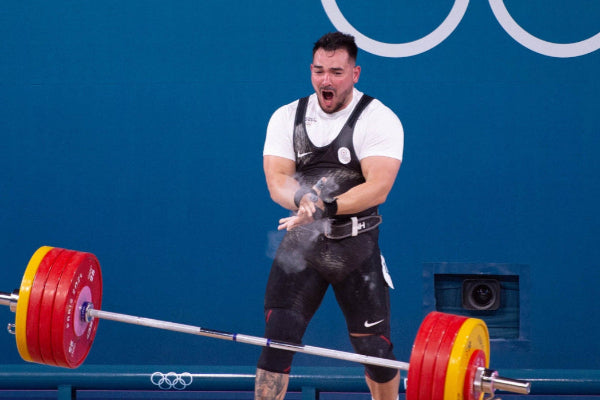 Weightlifter lifting a barbell with red weight plates in front of an Olympic logo.